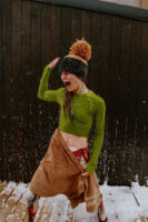 A woman standing in a dusting of snow on a wooden deck, in front of a black wood wall, poses in an expressive shock look after a wellness cold plunge and sauna session at Cedar and Stone, in a green crop top made of spandex shirt and brown towel skirt over leggings printed with flesh and bone.