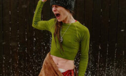 A woman standing in a dusting of snow on a wooden deck, in front of a black wood wall, poses in an expressive shock look after a wellness cold plunge and sauna session at Cedar and Stone, in a green crop top made of spandex shirt and brown towel skirt over leggings printed with flesh and bone.