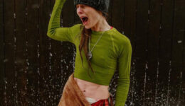 A woman standing in a dusting of snow on a wooden deck, in front of a black wood wall, poses in an expressive shock look after a wellness cold plunge and sauna session at Cedar and Stone, in a green crop top made of spandex shirt and brown towel skirt over leggings printed with flesh and bone.