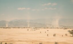 Wide horizon view of Black Rock City during burning man, small dots of people, bicycles, and dust torrnados with a washed out blue sky with small clouds above a dusty mountain scape