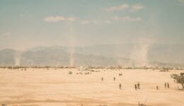 Wide horizon view of Black Rock City during burning man, small dots of people, bicycles, and dust torrnados with a washed out blue sky with small clouds above a dusty mountain scape