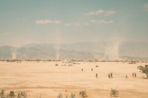 Wide horizon view of Black Rock City during burning man, small dots of people, bicycles, and dust torrnados with a washed out blue sky with small clouds above a dusty mountain scape